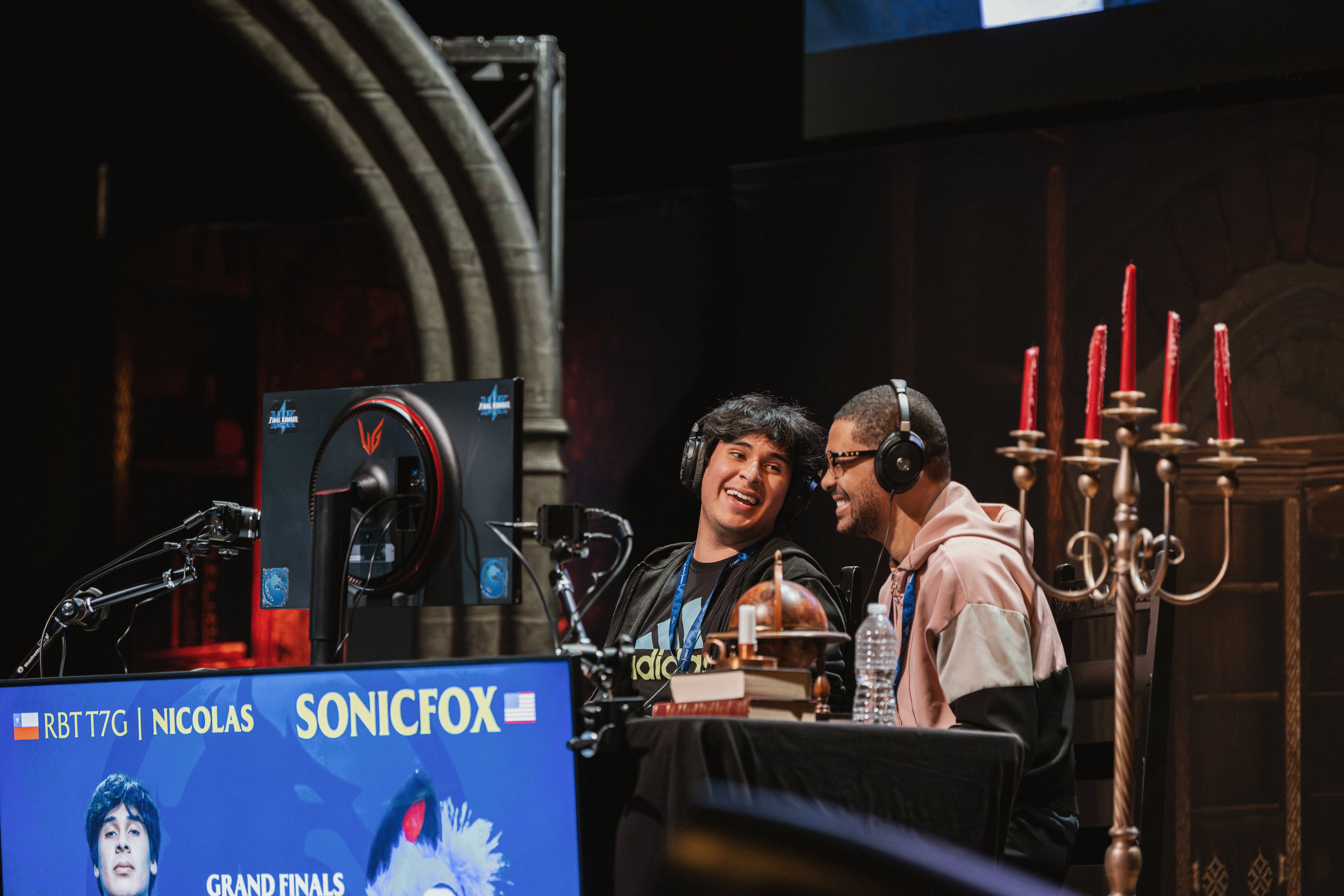 Two male esports commentators wearing headsets sit at a broadcast desk, smiling and looking at each other during a "Grand Finals" event, with a monitor displaying player names and a gothic-style candelabra in the background.