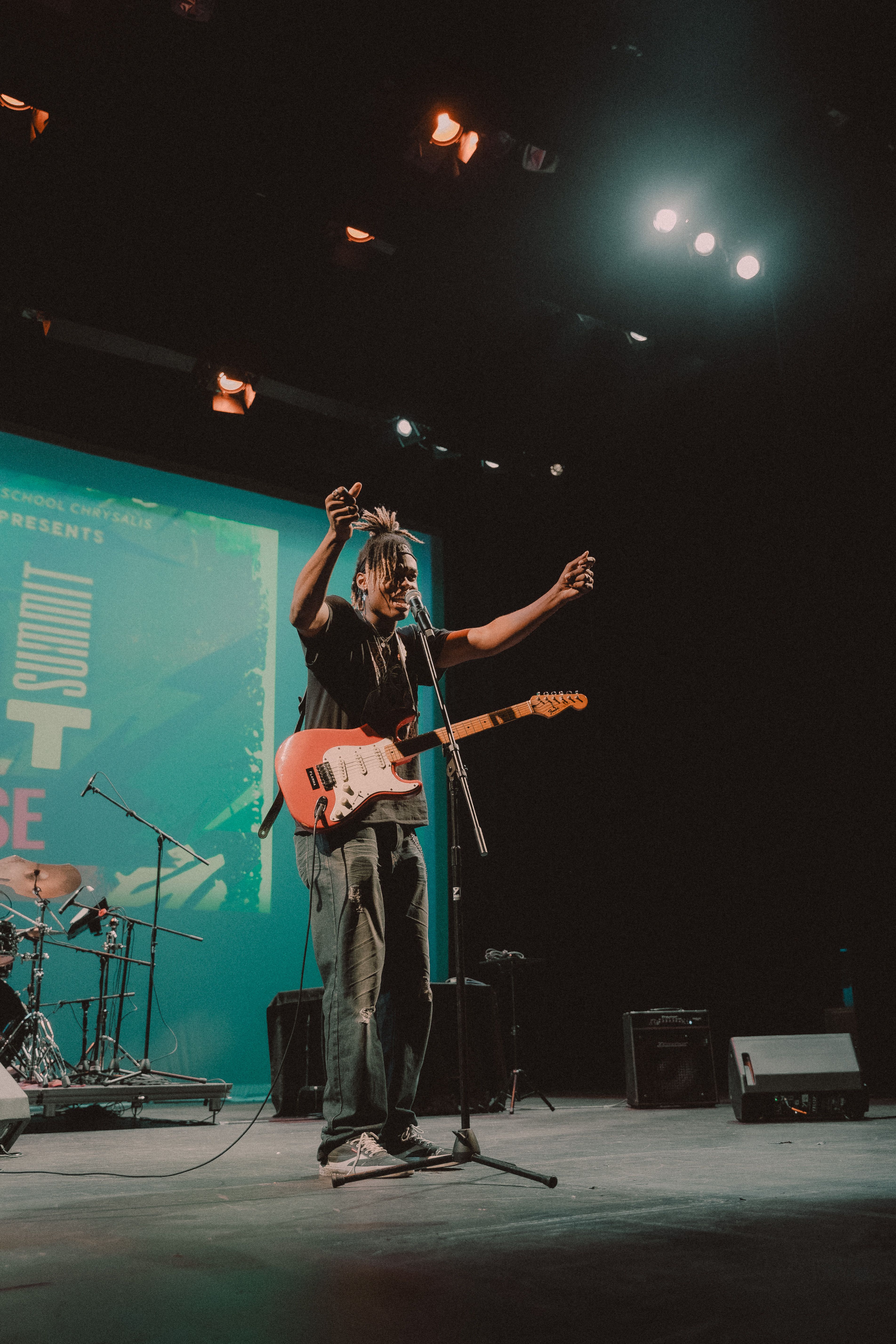 A musician with dreadlocks, wearing a black shirt and ripped jeans, stands on a stage with his arms raised, singing into a microphone while playing a red electric guitar, illuminated by stage lights against a large blue-green backdrop.