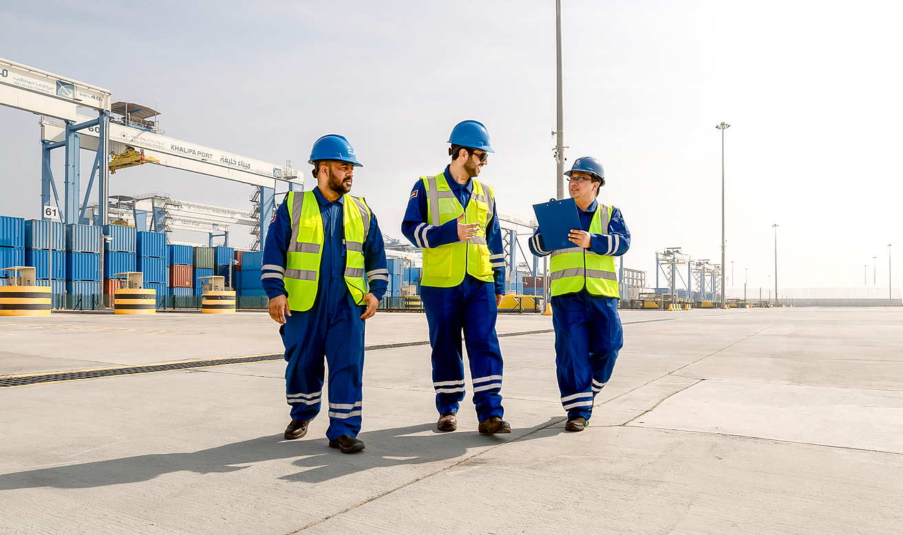 Three workers in safety gear are conversing as they walk together across a large empty yard at a port.
