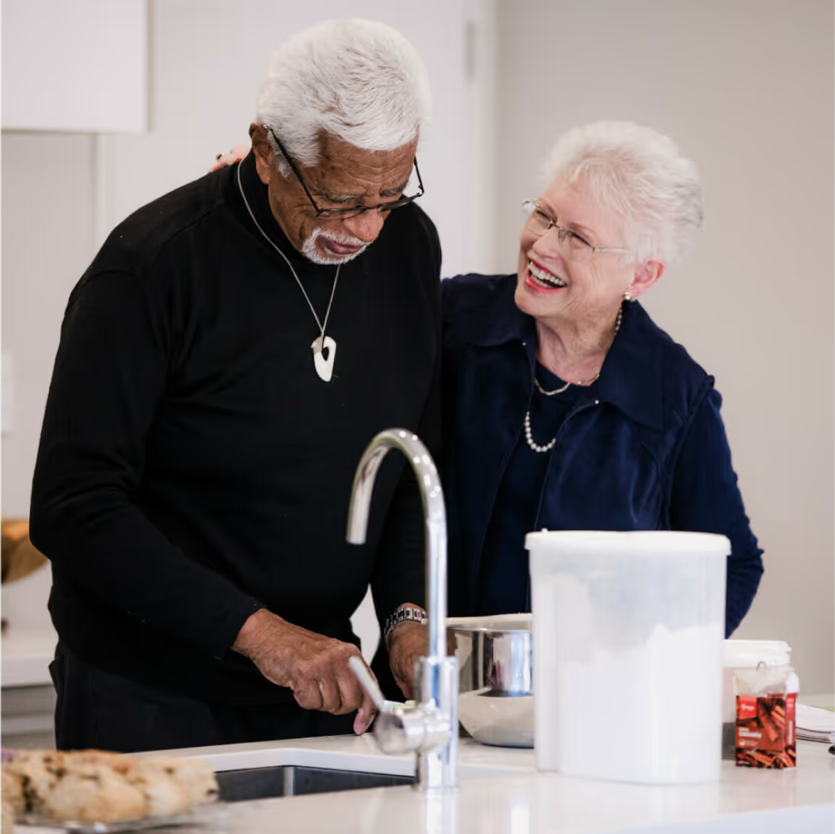 An older couple, both wearing glasses, interact while they're standing in a kitchen
