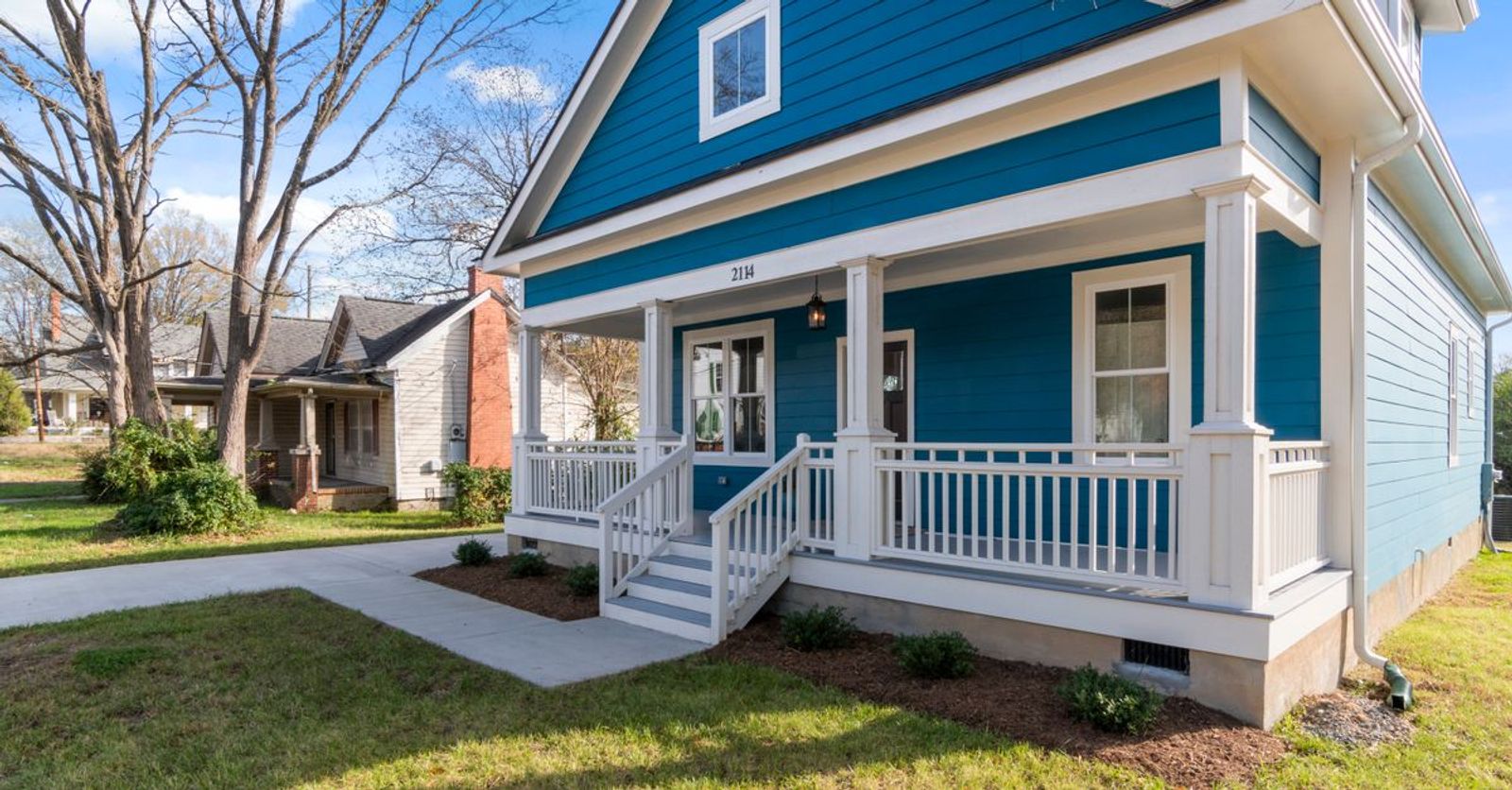 A cozy blue suburban house with a white front porch and lush surroundings on a clear day.