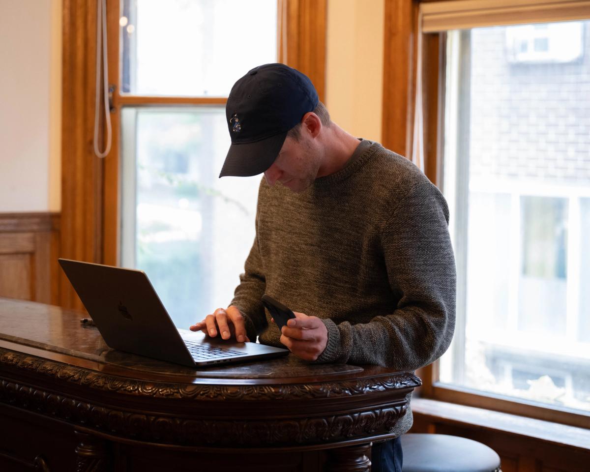 Cohley co-founder Erik Graber in a baseball cap, working on a laptop while holding a phone at a wooden counter during a company offsite in Milwaukee