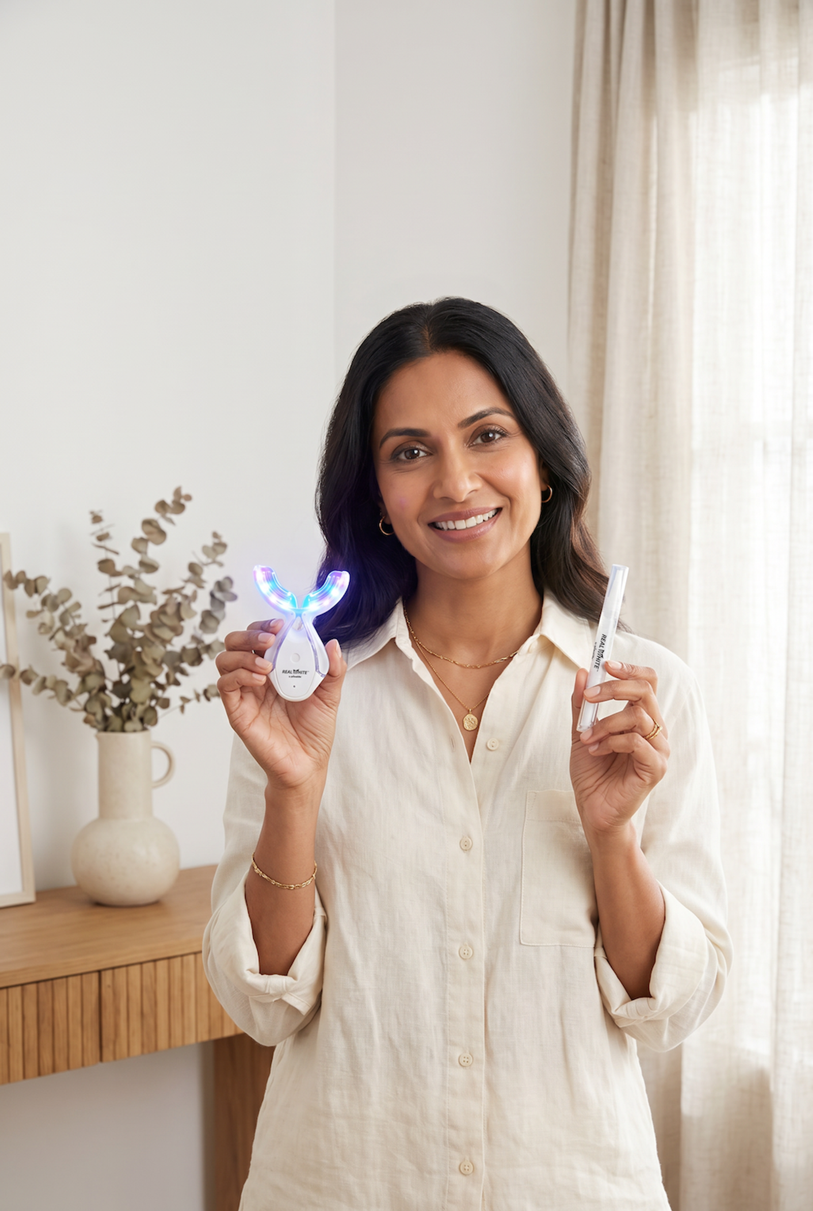 Woman smiling while holding a Real White LED teeth whitening device and a whitening pen