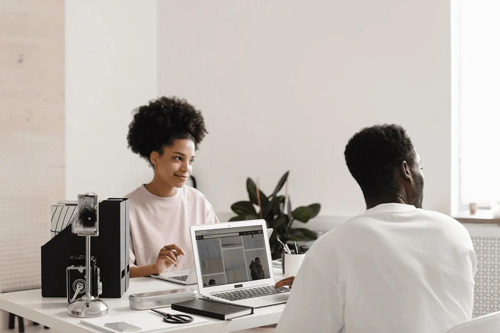 Two colleagues talking across a desk with laptops in a bright office