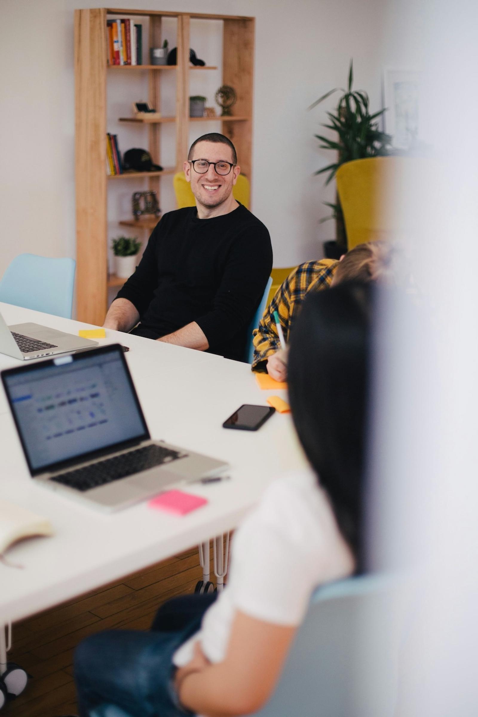 Smiling man at an office meeting table with laptops and colleagues