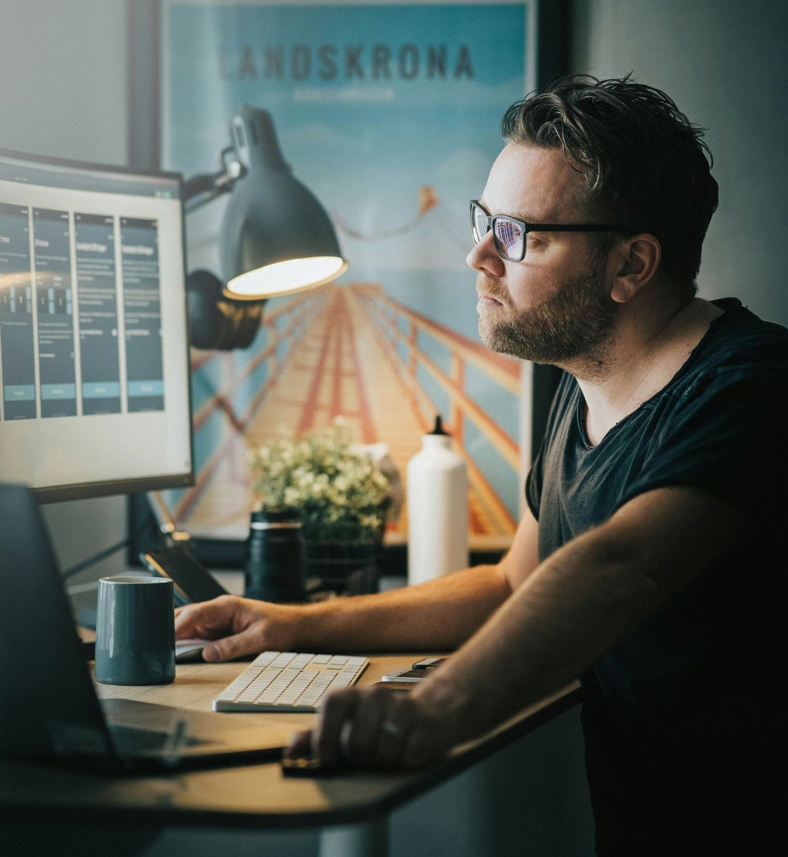 Man with glasses reviewing analytics dashboards on two monitors at a warm-lit desk