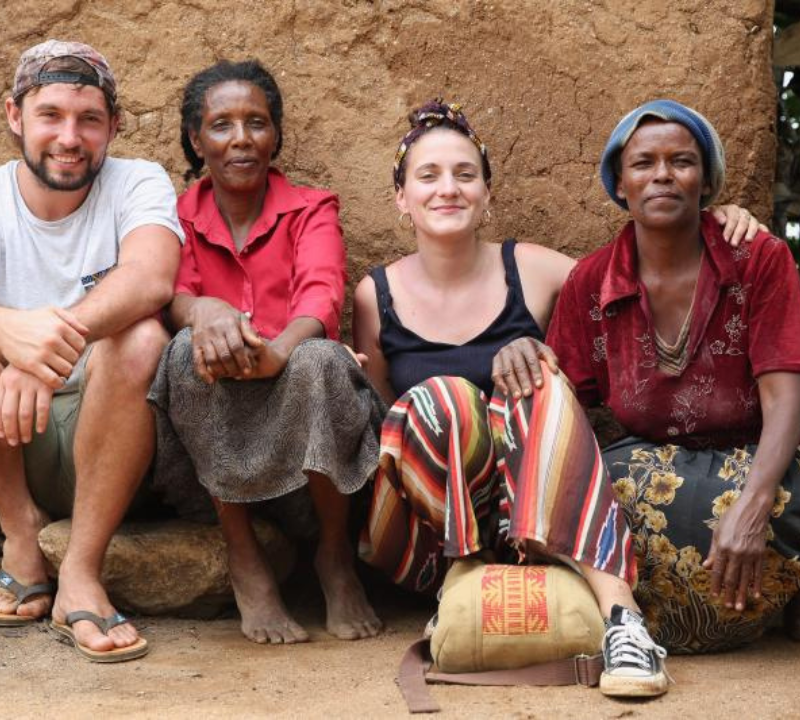 Four people, two white and two African women, sit closely together in front of an earthen wall. One white woman has her arm around an African woman.