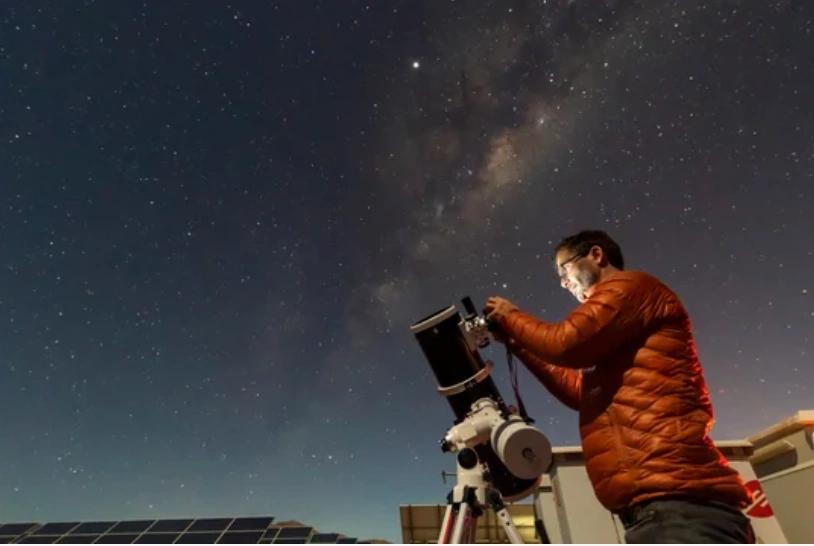 A person adjusts a telescope under a starry night sky with the Milky Way visible, above solar panels.