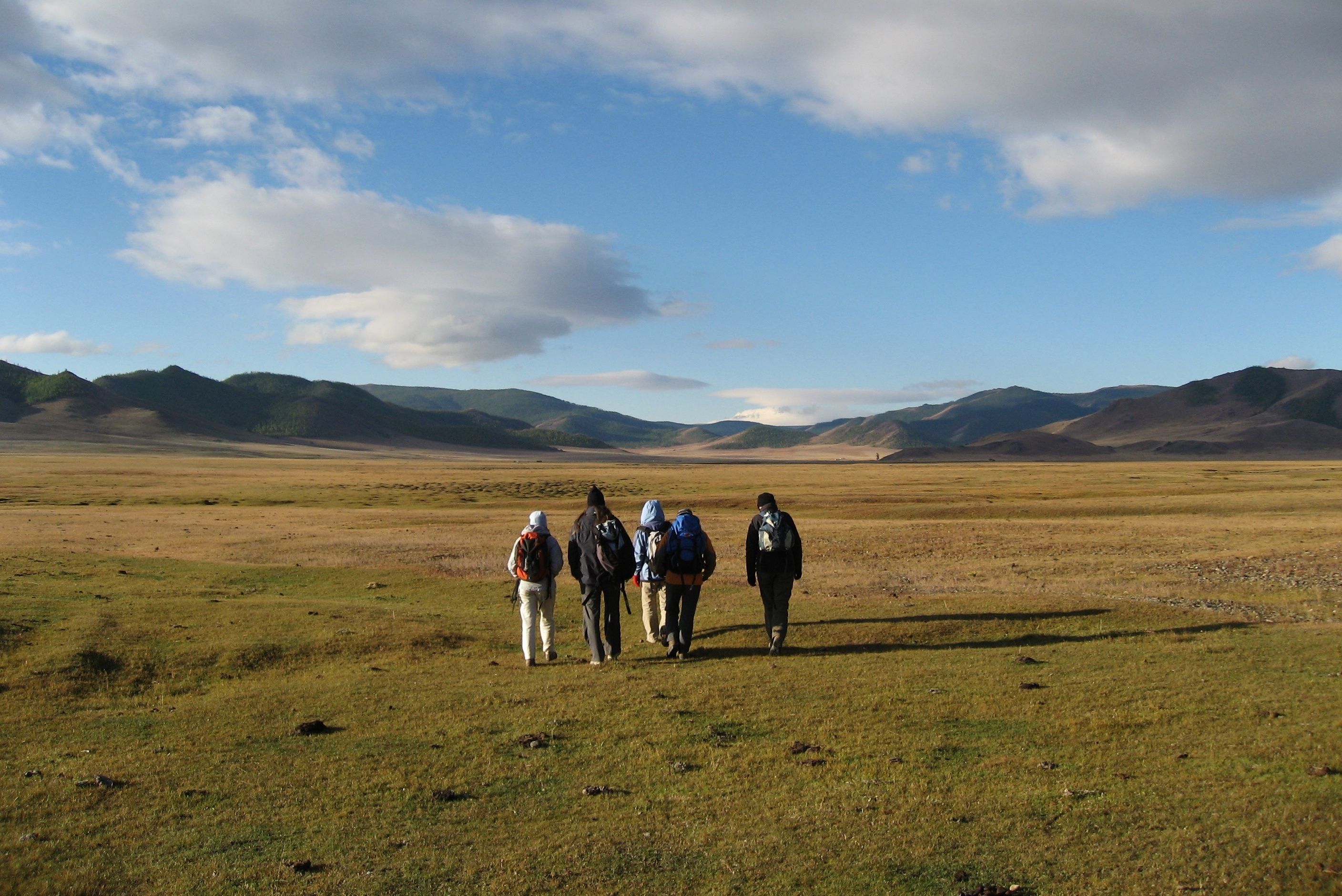 Four hikers walk across a wide grassy plain towards distant mountains.