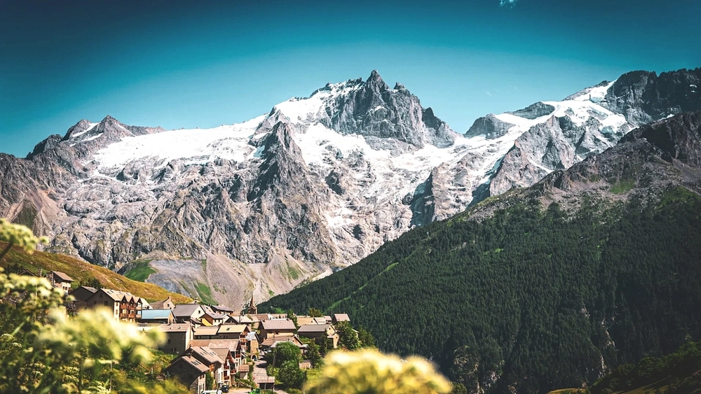 A small village sits beneath towering snow-capped mountains and forested slopes.