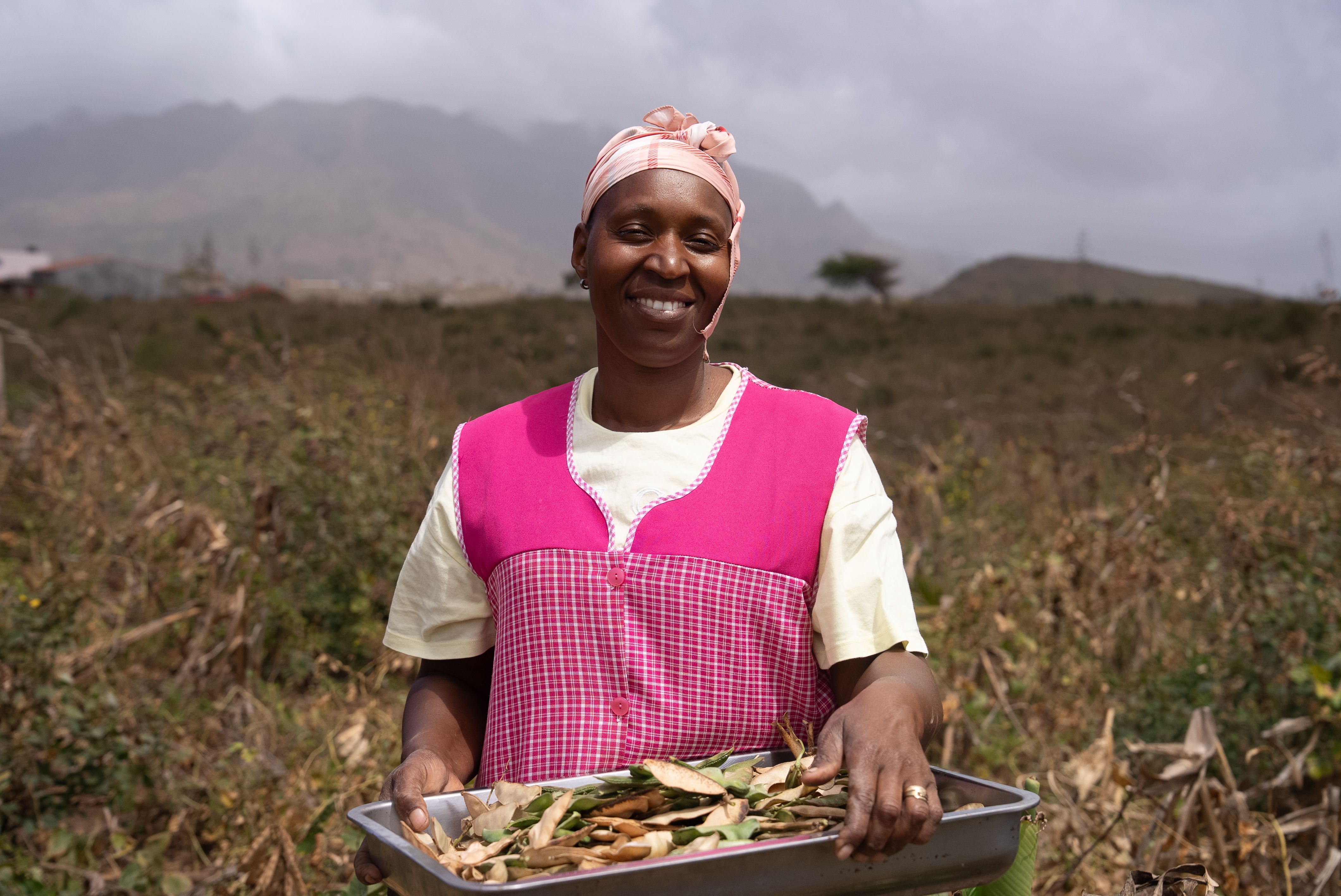 A smiling Black woman in a pink apron holds a tray of vegetable scraps in a field, with mountains in the background.