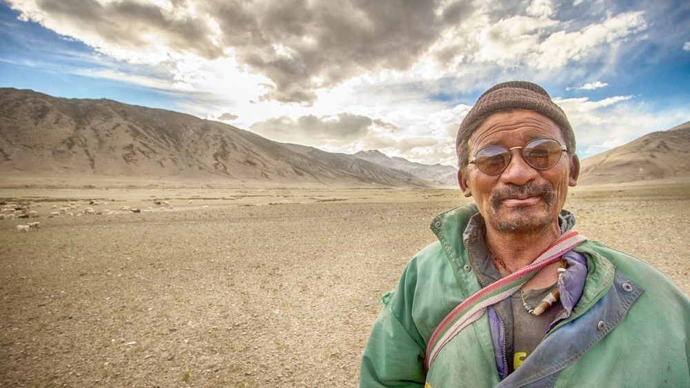 A smiling man in sunglasses and a beanie stands in a wide, dry mountain valley under a cloudy sky.