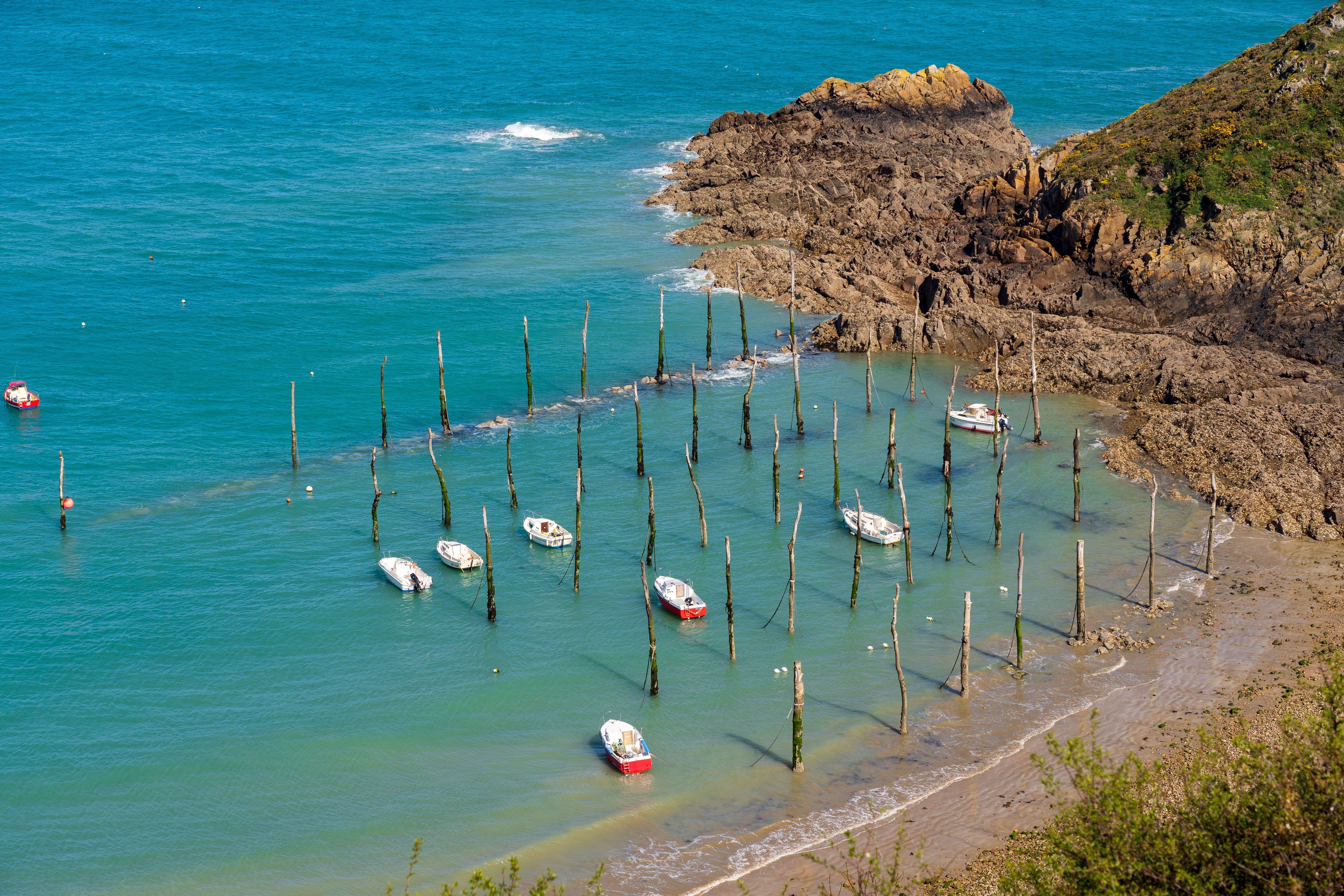 Small boats moored to wooden poles in a turquoise bay with a sandy beach and rocky cliffs.