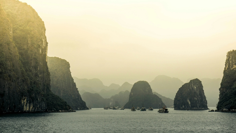 A hazy bay scene with numerous boats floating between towering limestone karsts under a golden sky.