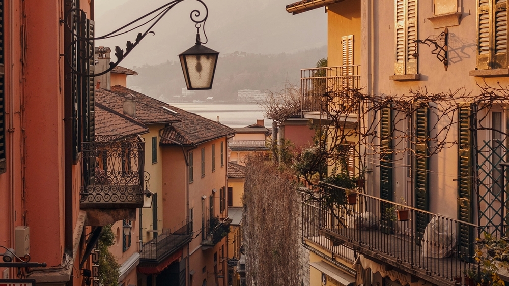 A steep cobblestone street descends between colorful buildings to a hazy lake and mountains.