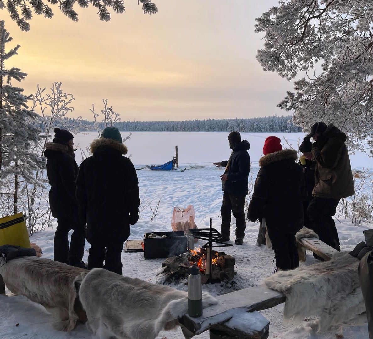 A group of people gather around a campfire in a snowy forest overlooking a frozen lake at sunset.