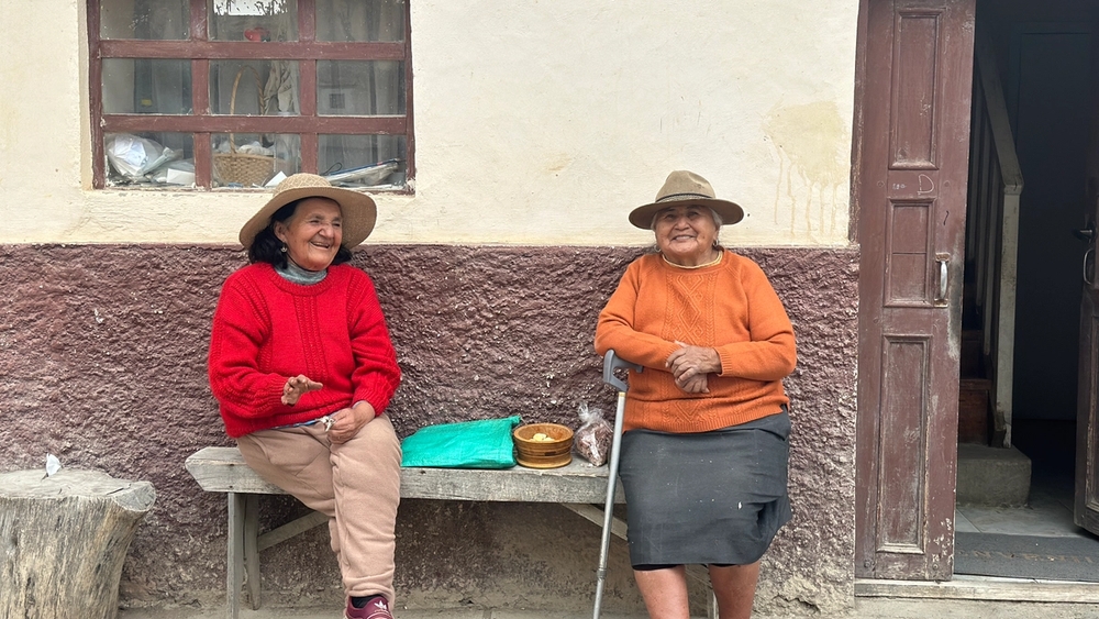 Two smiling older women in hats sit on a wooden bench outside a building.