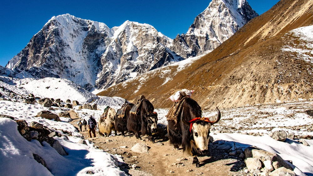 A snow-covered mountain peak rises behind a rocky hill draped with colorful Tibetan prayer flags, with a few people visible.
