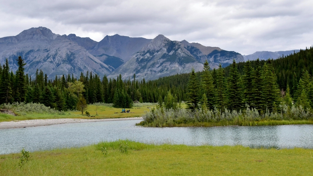 A calm river flows past evergreen forests with grey mountains rising in the background under a cloudy sky.