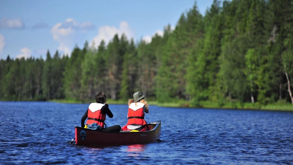 Two people in red life vests paddle a canoe on a lake with a forest background.