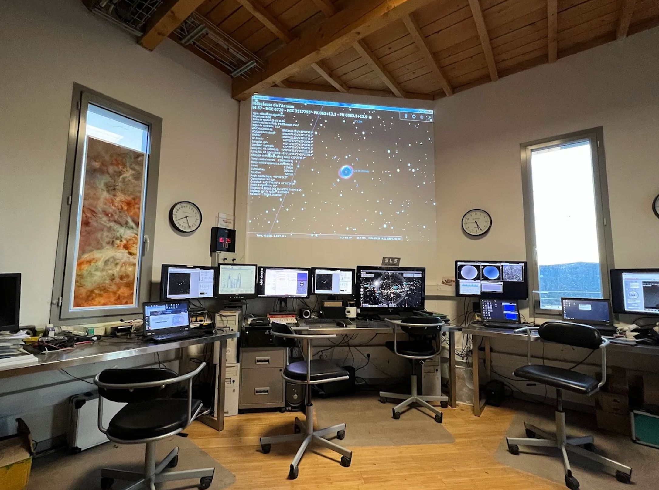 A control room with multiple computer monitors, a large projected star map on the wall, and a colorful nebula graphic in a window.