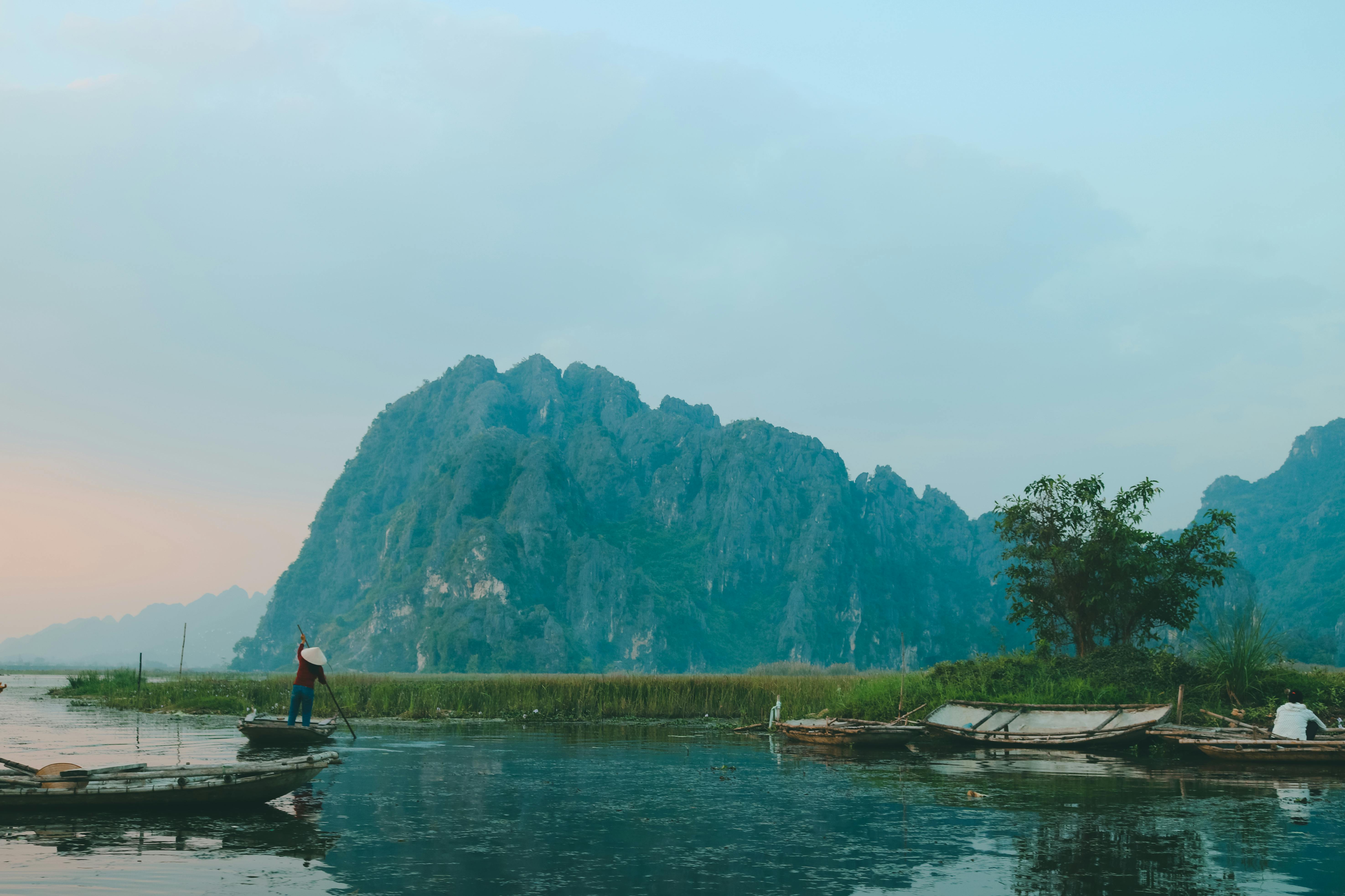 A person in a conical hat paddles a boat on a river with lush, rocky mountains in the background.