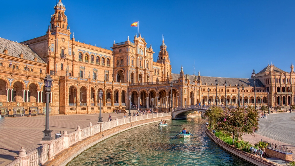 Ornate, orange-toned Plaza de España in Seville, with a canal, rowboats, and people under a bright blue sky.