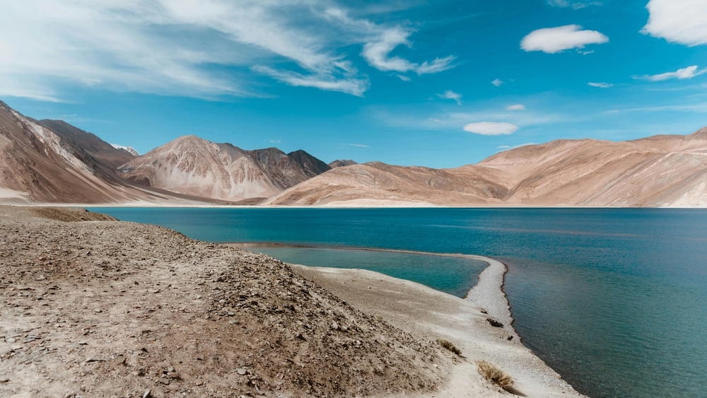 A bright blue lake framed by arid, tan mountains under a blue sky with scattered white clouds. A narrow sandy spit extends into the water.