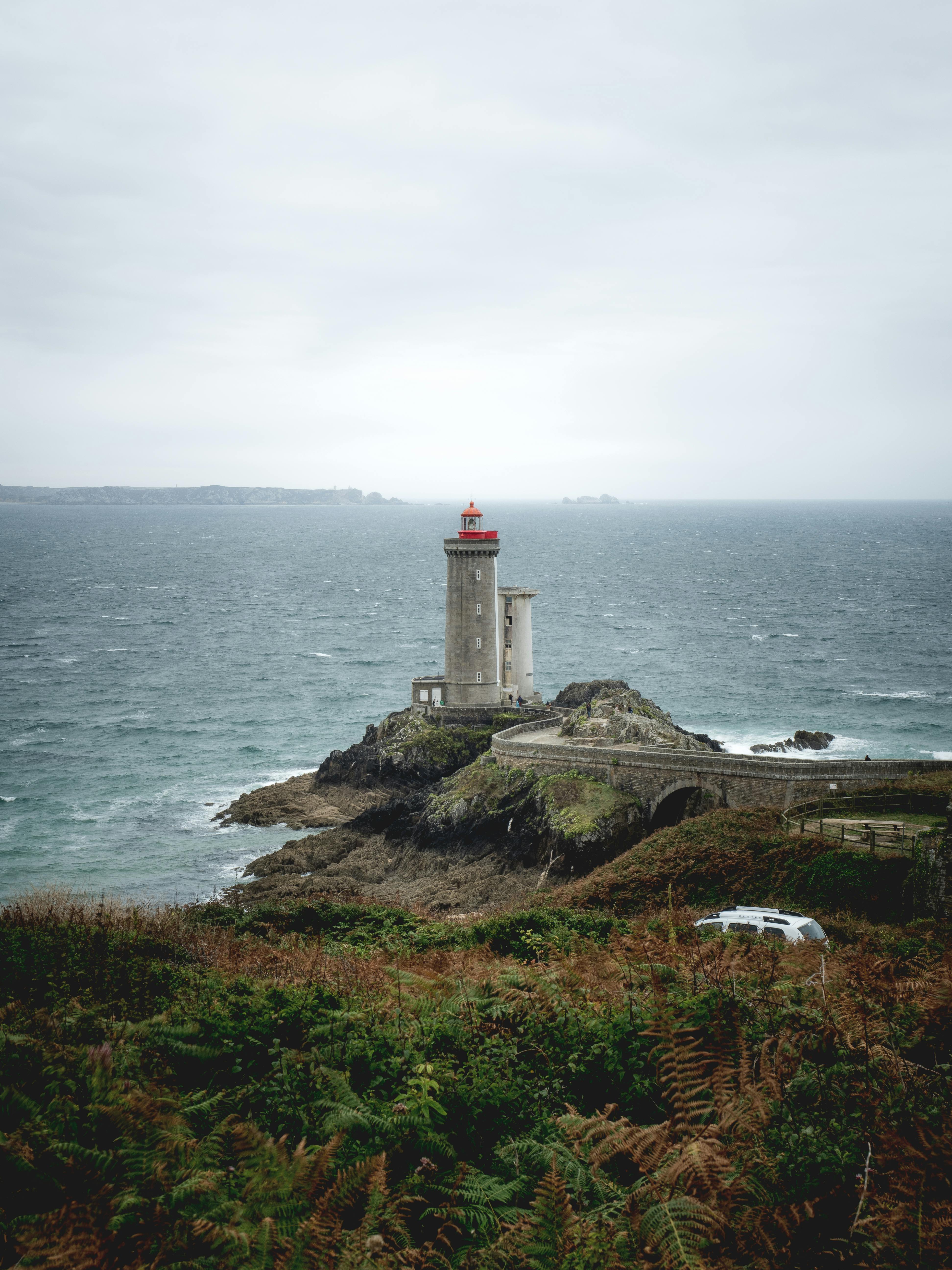 A stone lighthouse with a red cap stands on a rocky islet, connected by an arched stone bridge to a fern-covered coast, overlooking a grey sea under a cloudy sky.