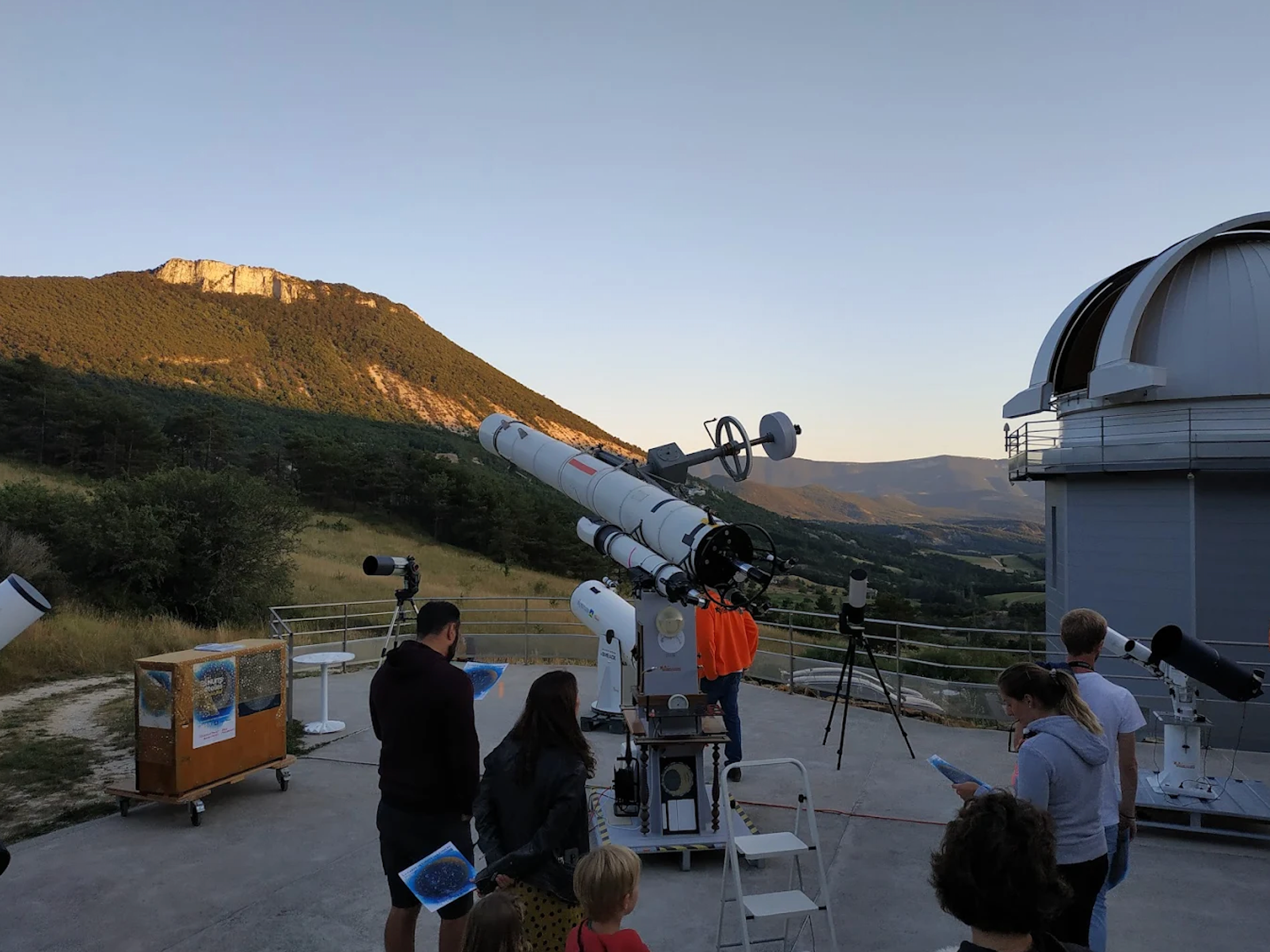 People at an outdoor observatory with multiple telescopes, a dome, and mountains at dusk.