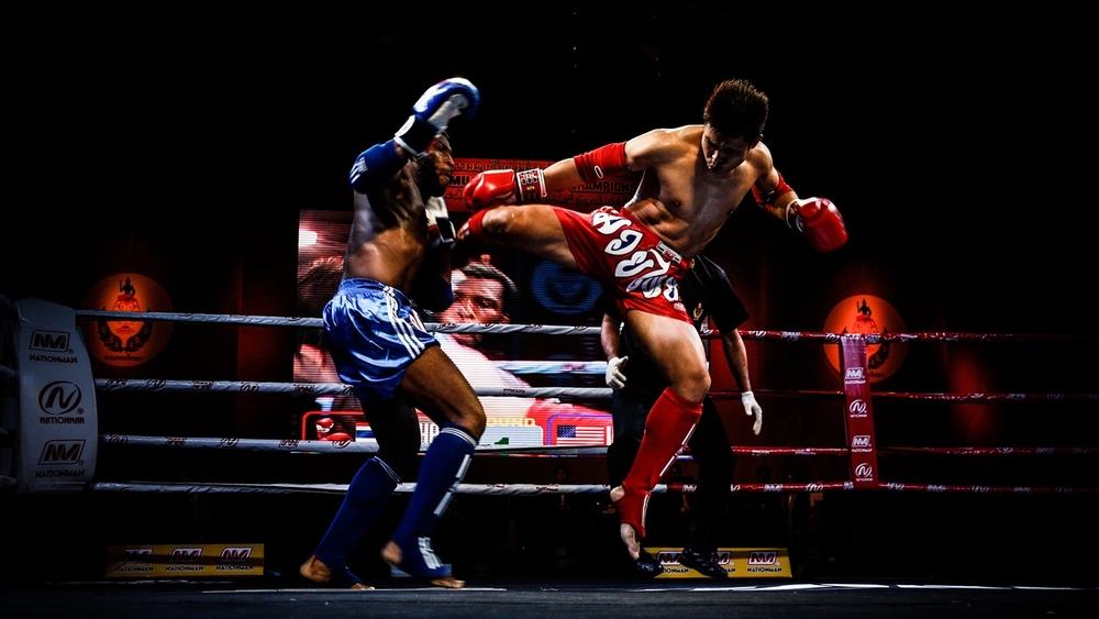 A Muay Thai fighter in red shorts mid-air kicks an opponent in blue shorts in a boxing ring.