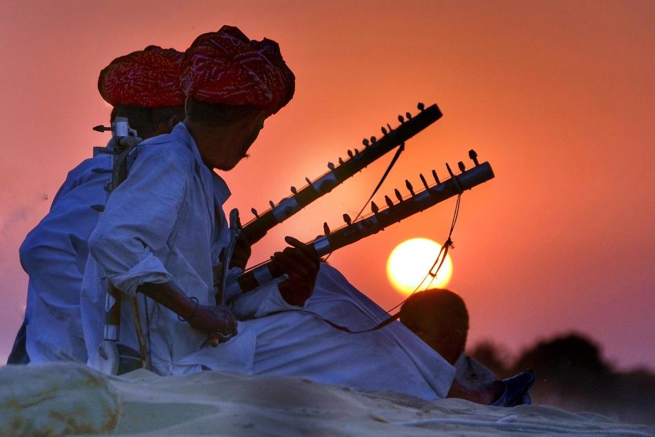 Two turbaned musicians play instruments on sand dunes at sunset.