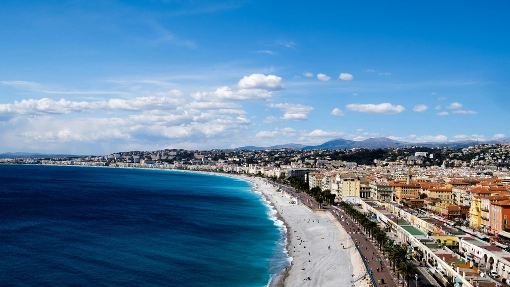 Panoramic view of the vibrant Nice coastline with a deep blue ocean, white pebble beach, promenade, and colorful buildings under a clear sky.