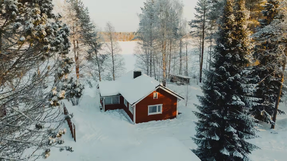 A small red cabin in a snow-covered forest overlooking a frozen lake.