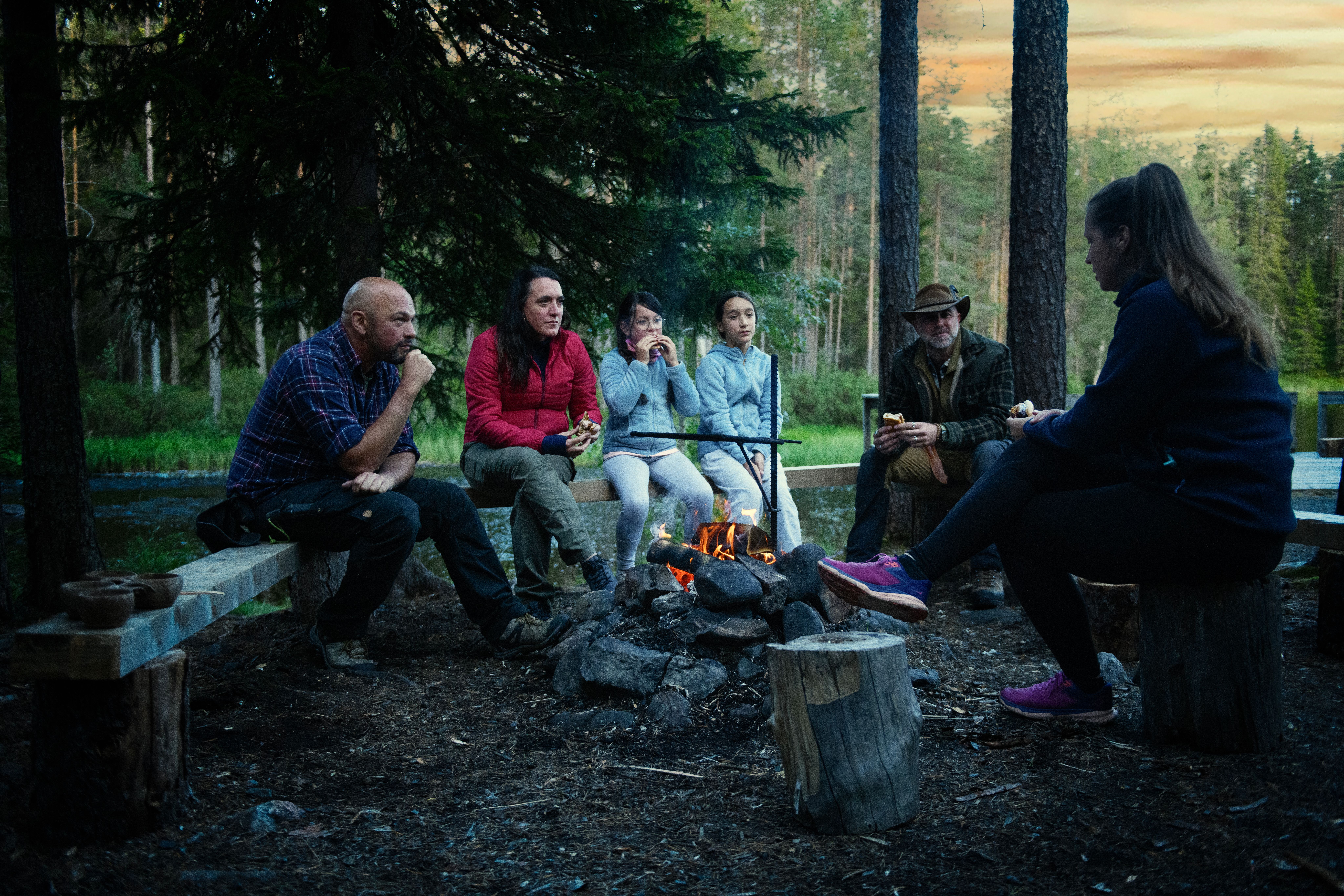 Six people sit around a campfire in a forest at dusk, next to a lake.