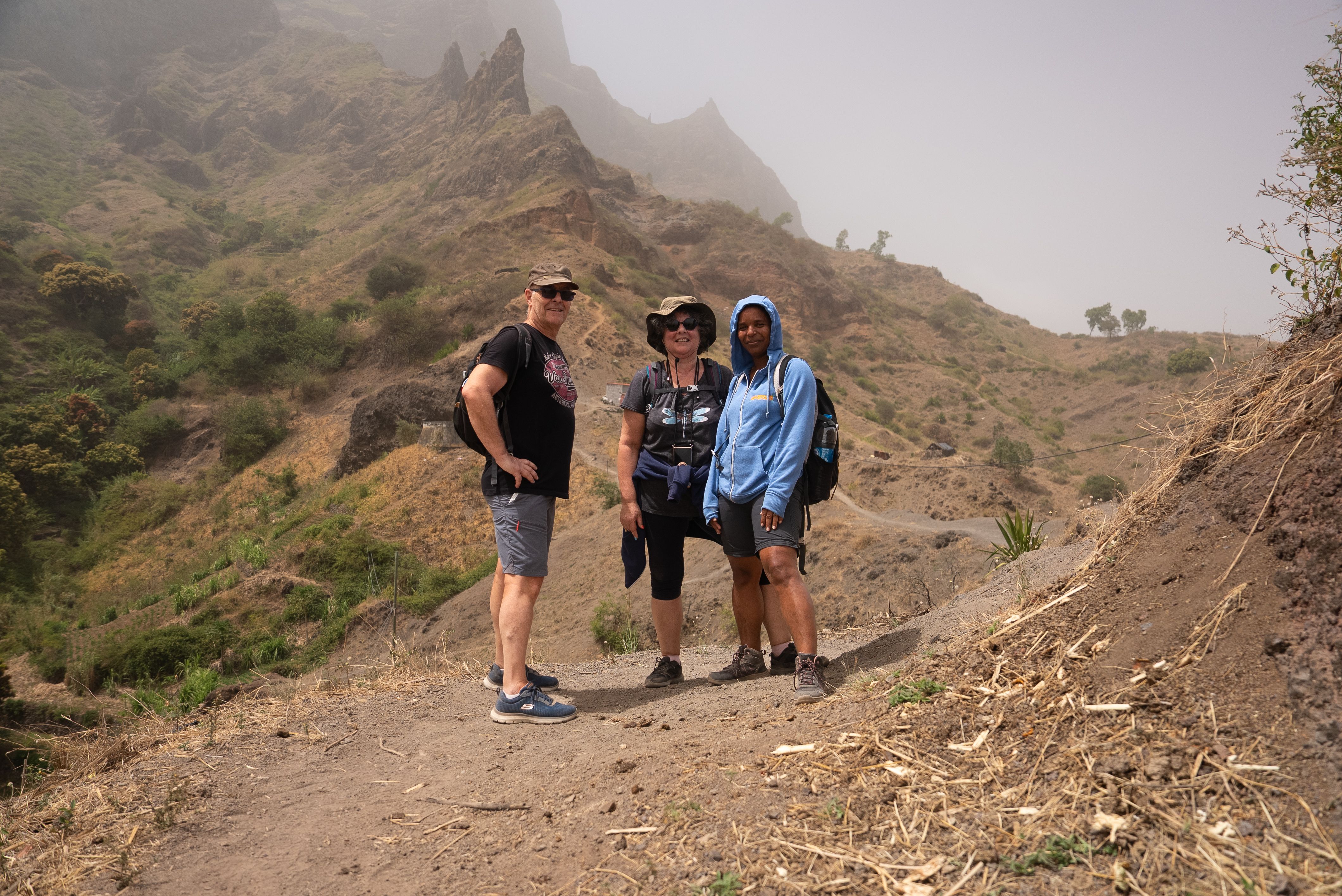 Three hikers stand on a dirt path in a hazy, mountainous landscape.