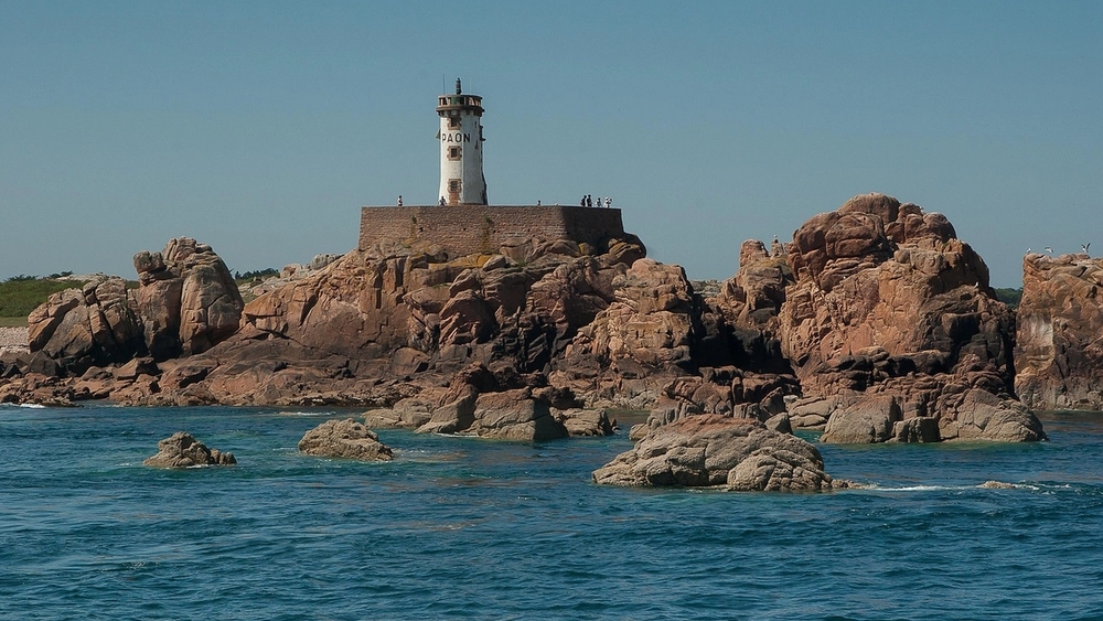 A white lighthouse on a rugged reddish-brown rocky island, surrounded by blue water under a clear sky.