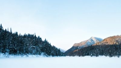 A snowy winter landscape with a frozen expanse, evergreen forests, and distant mountains with sunlit peaks.