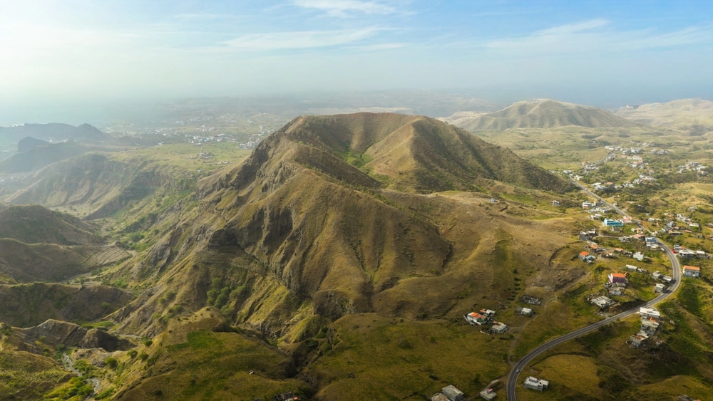 A white lighthouse on a rocky peninsula at sunset, with the sun low over the ocean and a distant coastal town.