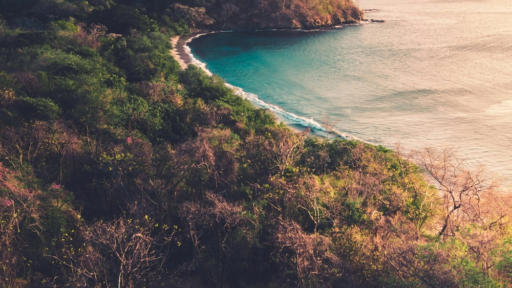 Aerial view of a tropical bay with turquoise water, a sandy beach, and lush hills under a warm sky.