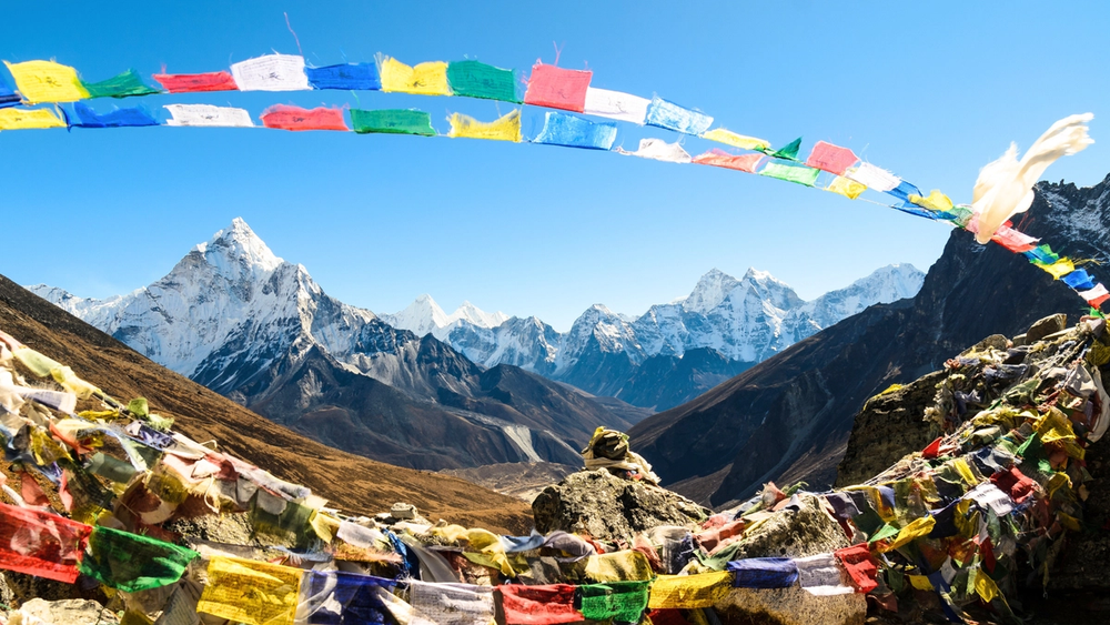 Colorful prayer flags with Ama Dablam and snow-capped Himalayan peaks under a clear blue sky.