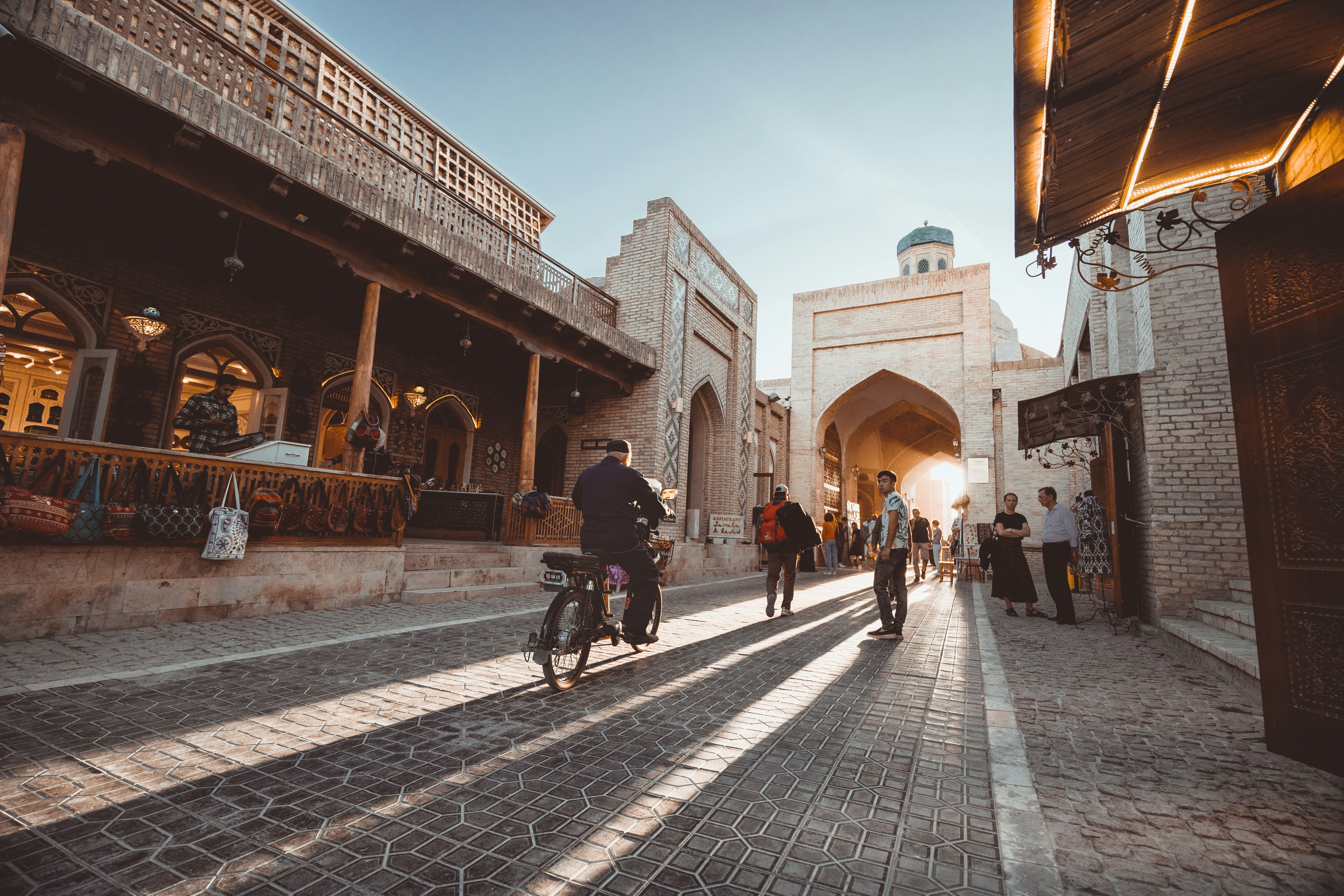 Historic city street with traditional brick buildings, archways, and a dome under golden sunlight, casting long shadows from people and a moped.