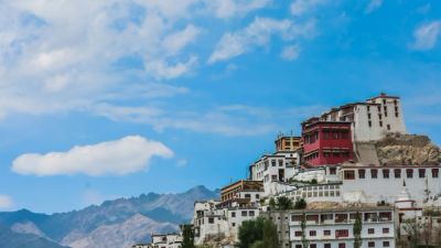 A white and red tiered monastery complex built on a rocky hill, with distant mountains under a blue sky.