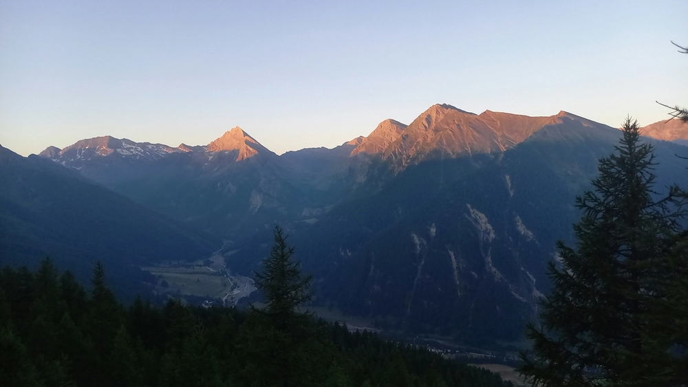 Mountain peaks glow with golden light above a river valley, framed by dark foreground trees.