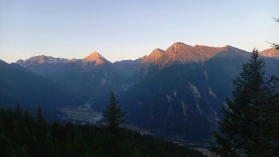 Mountain peaks glow with golden light above a river valley, framed by dark foreground trees.