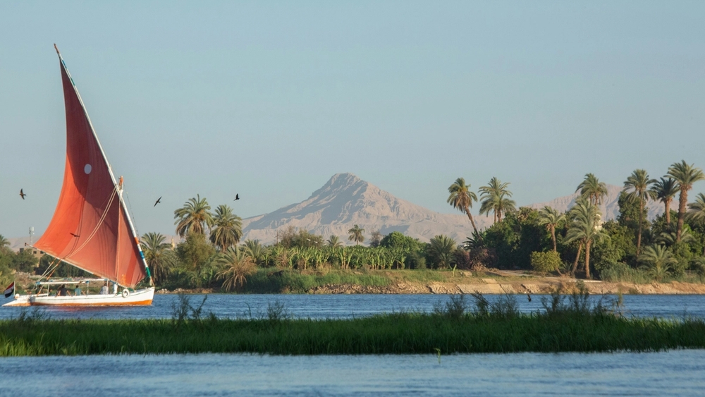 A felucca with an orange sail glides on a river lined with palm trees, under a clear sky with mountains in the distance.