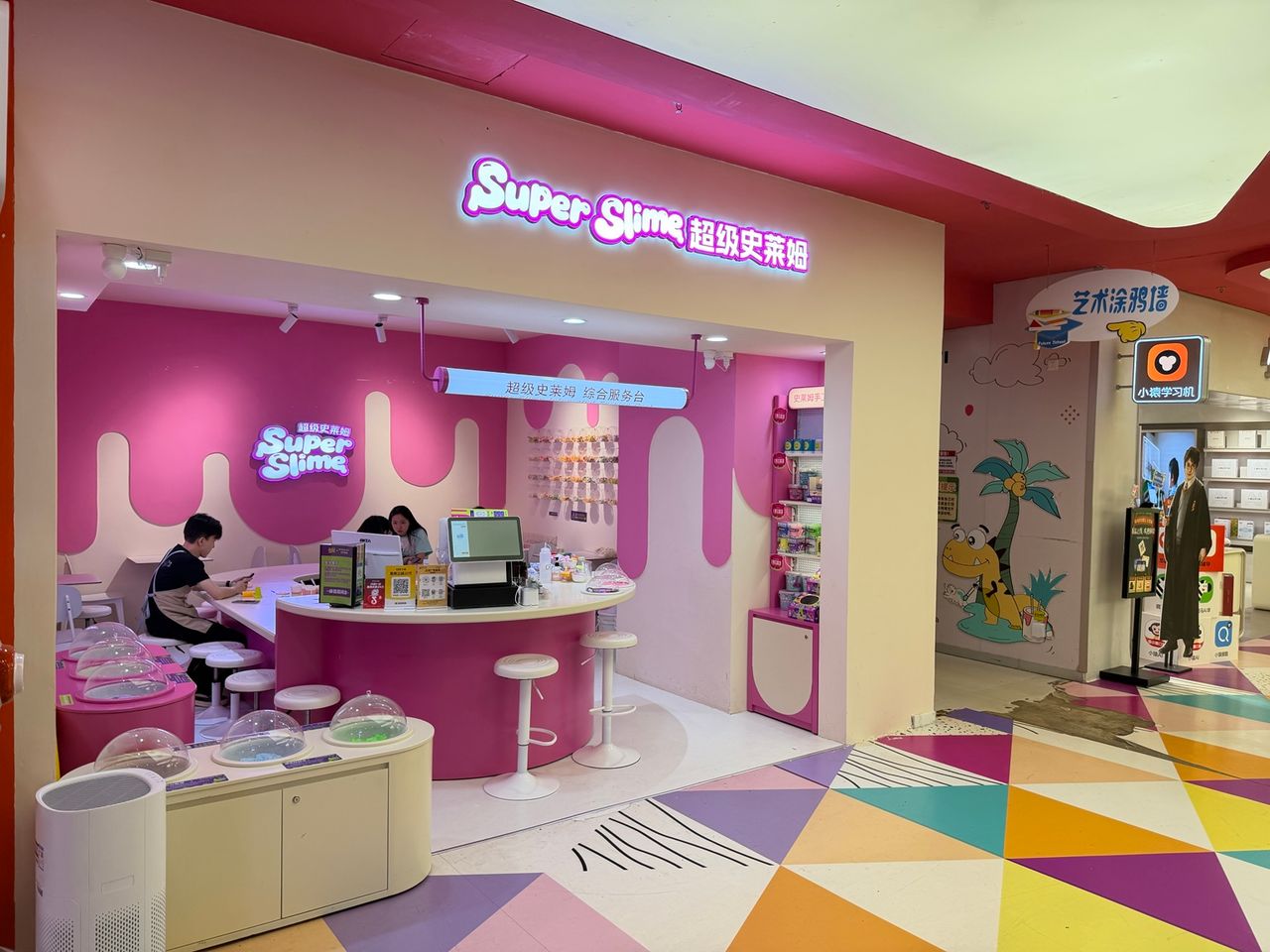 Photo terrain STORIMITIK montrant l’entrée et l’intérieur d’une boutique Super Slime en Chine, avec tables, tabourets et comptoir rose.