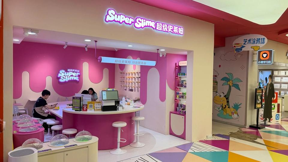 Photo terrain STORIMITIK montrant l’entrée et l’intérieur d’une boutique Super Slime en Chine, avec tables, tabourets et comptoir rose.