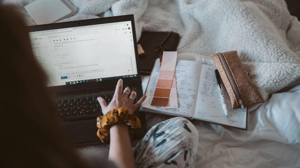 Une femme travaille depuis son domicile sur un ordinateur portable, entourée de notes et de fournitures.