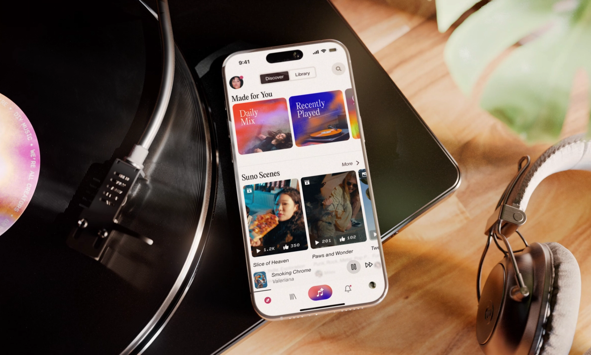 A smartphone rests on a spinning vinyl record. The screen displays the Suno app with sections like “Daily Mix,” “Recently Played,” and “Suno Scenes.” A pair of headphones and plant sits nearby on the wooden surface.